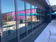 Dark-framed folding glass doors beside a patio, reflecting turquoise umbrellas and a colorful mural.