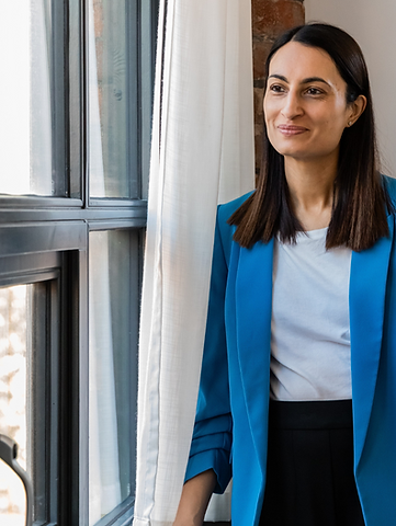Anita Patel standing by a window in a blue blazer, looking thoughtful and composed in a professional setting