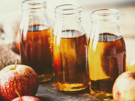 Three glass bottles of amber apple cider vinegar sit on a table with red apples around. Warm, rustic atmosphere with soft lighting.