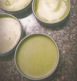 Four round salve tins filled with green salve on a speckled countertop surface. The salves have smooth textures and varying shades of green.