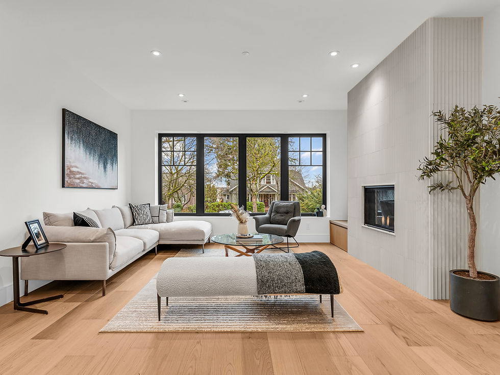 Vancouver Renovation Guide. Modern living room with beige sectional, gray chair, fireplace, plant, and large window view of trees and houses.