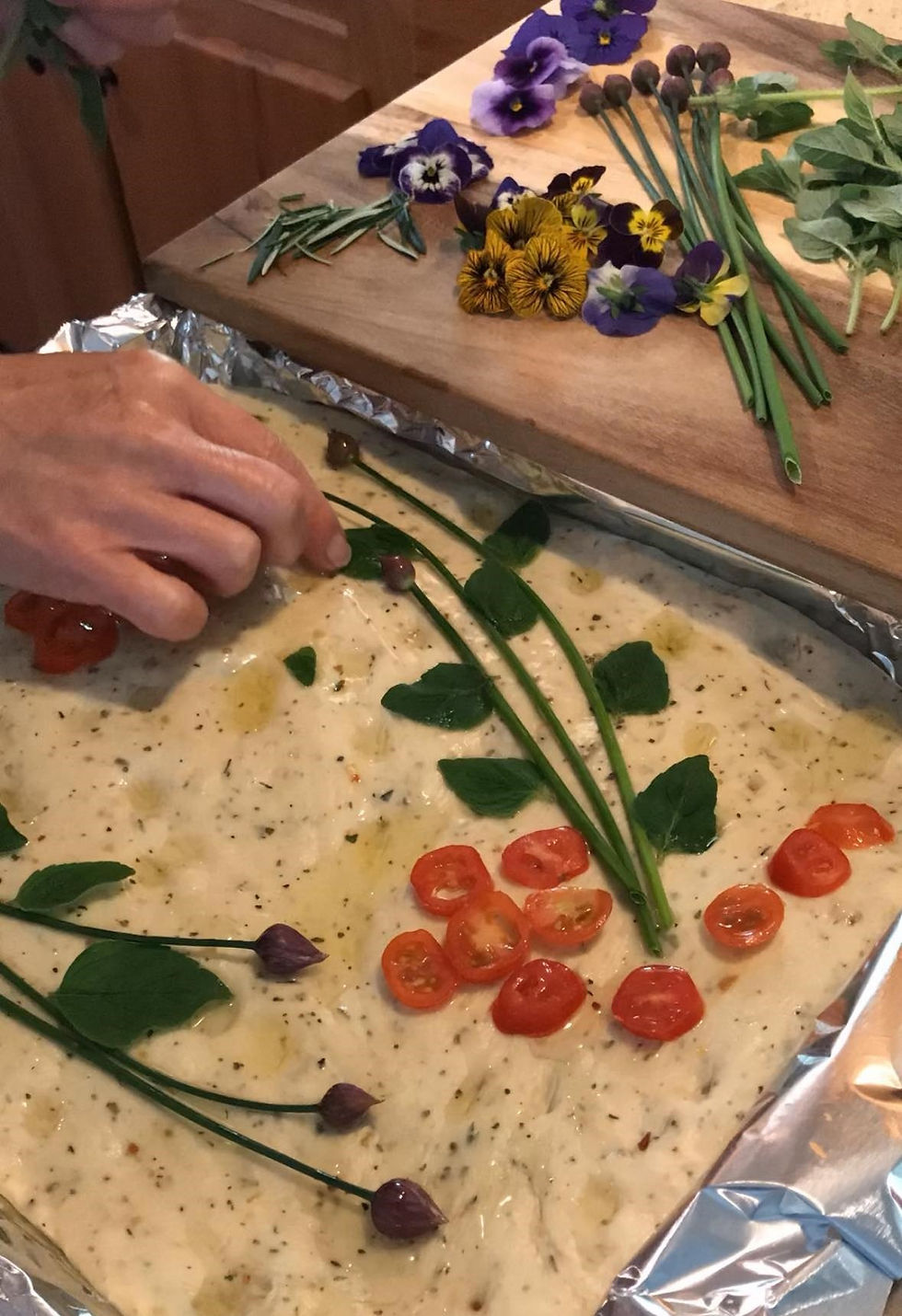 A hand decorates focaccia dough with cherry tomatoes and herbs. Colorful flowers and greens rest on a wooden board nearby.