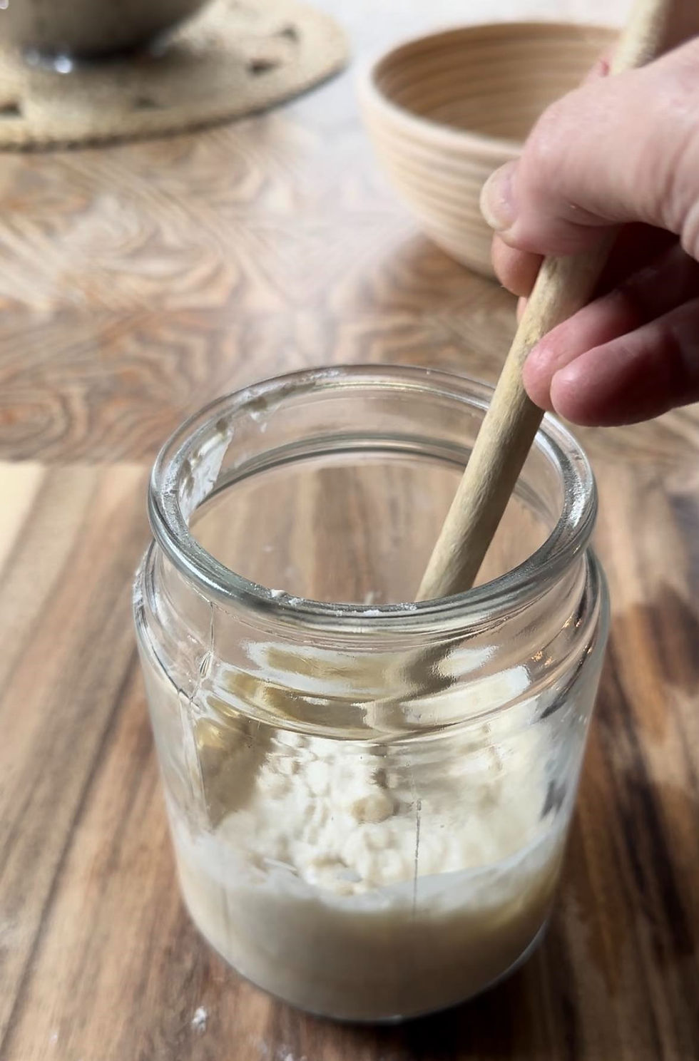 Hand stirs flour in a glass jar of sourdough starter with a wooden spoon on a patterned wooden table. A woven bowl is blurred in the background.
