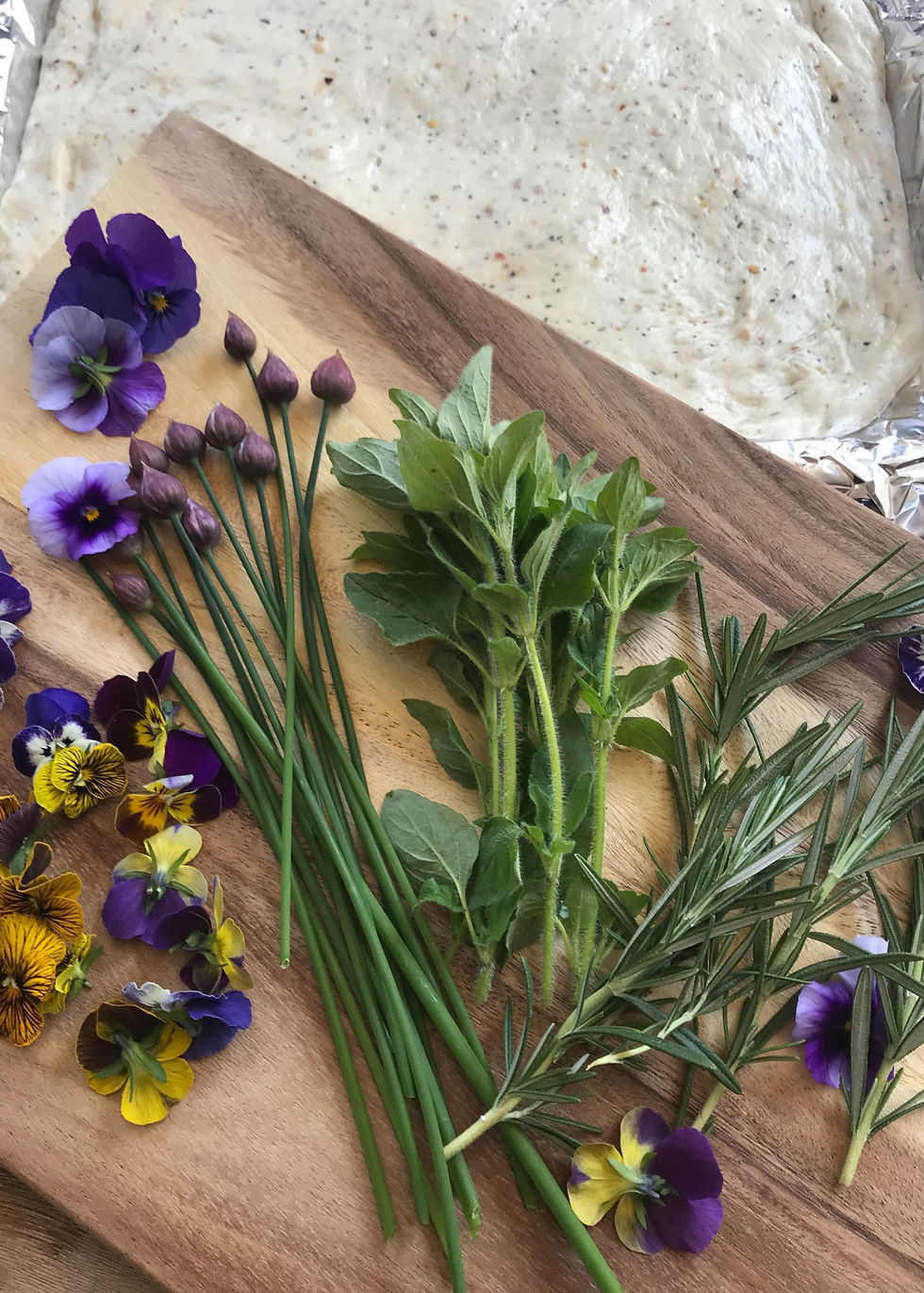 Herbs and edible flowers, like rosemary and pansies, on a wooden board. Dough in the background, with a fresh and colorful vibe.