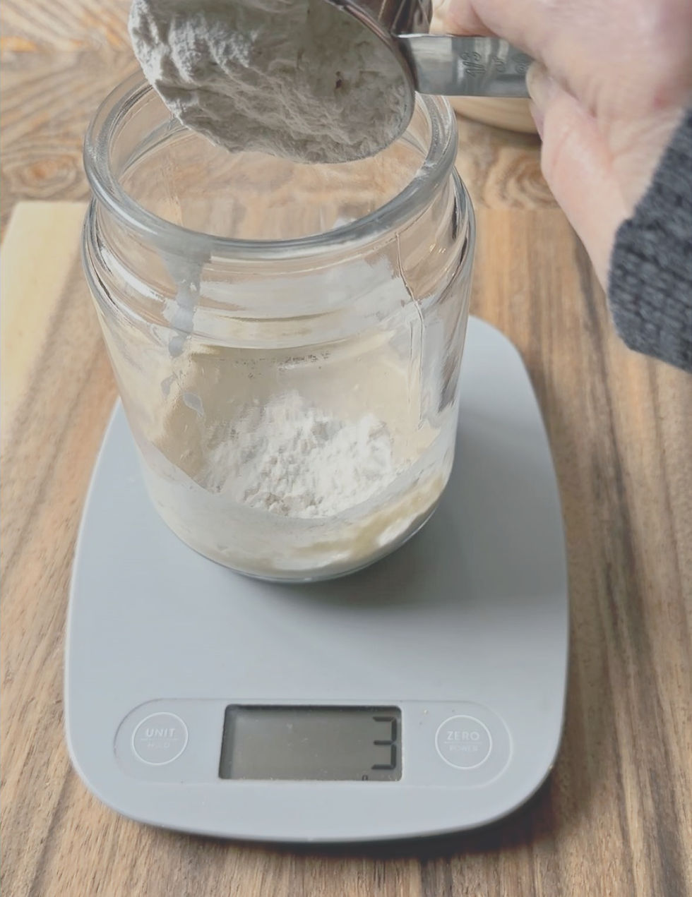 Hand pours flour from a cup into a jar on a digital scale reading "3" grams. The scene is set on a wooden surface.