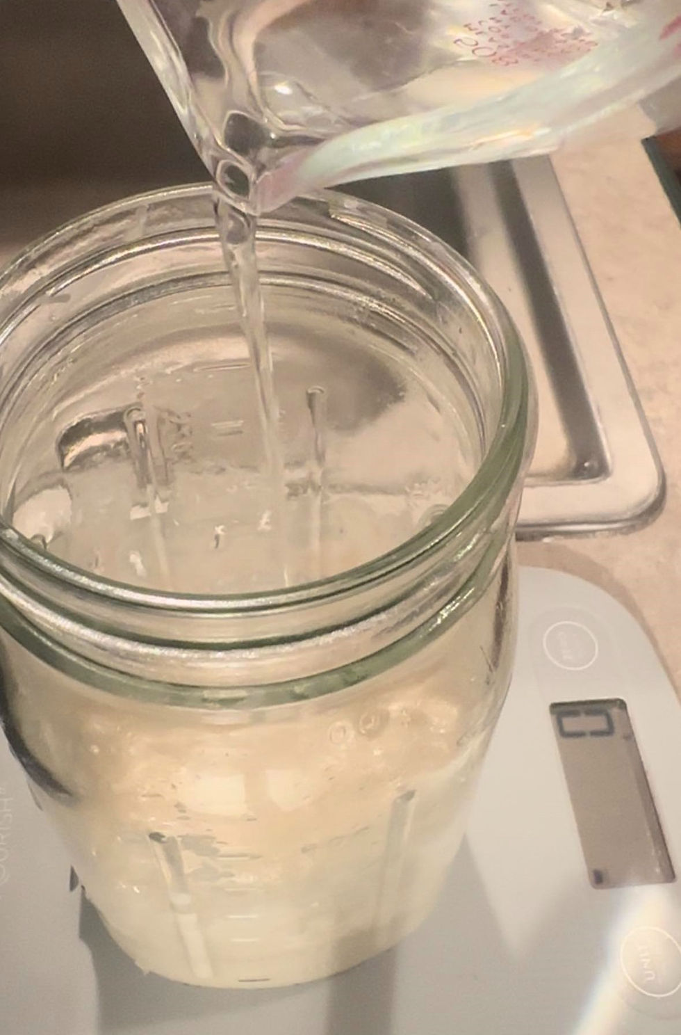 Water is being poured from a glass pitcher into a clear jar with sourdough starter on a digital scale. The setting is a neutral-colored kitchen counter.