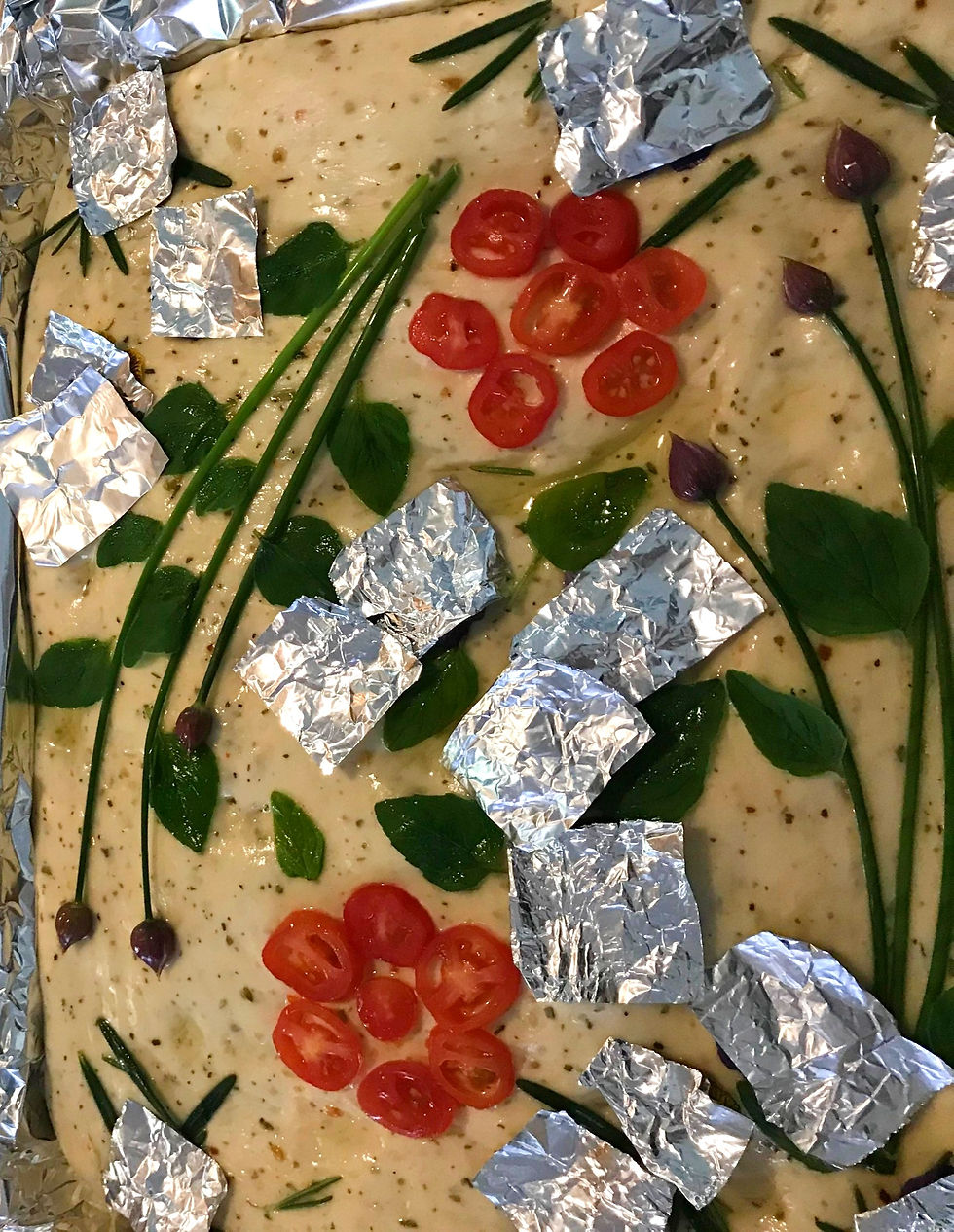 Unbaked focaccia dough decorated with cherry tomatoes, herbs, and foil squares, arranged on a baking tray. Colorful and creative.
