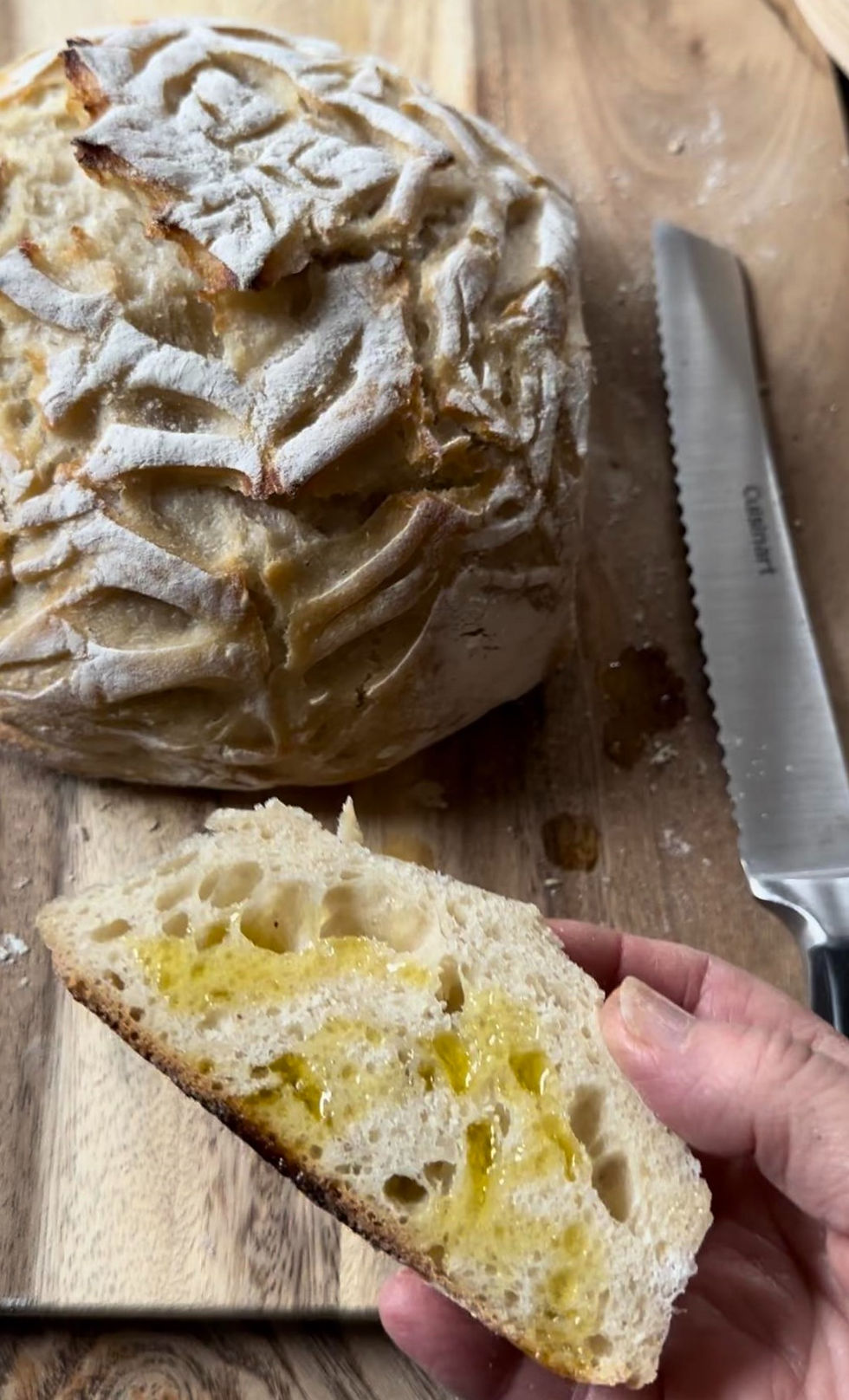 A hand holds a slice of sourdough bread with olive oil, next to a round loaf on a wooden board. A serrated knife is visible. Rustic scene.