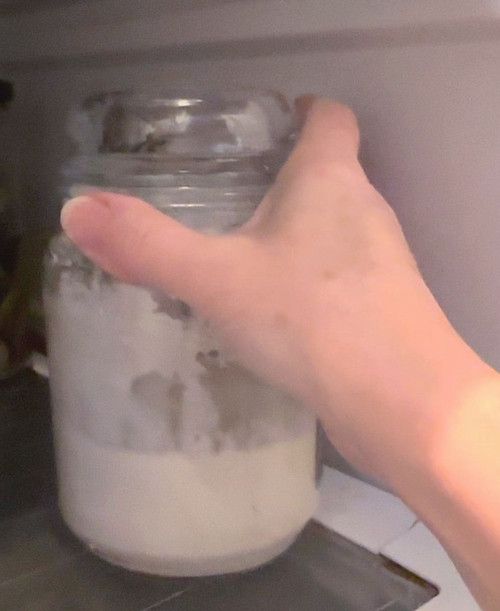 Hand placing a jar with white substance in a fridge. The setting is dimly lit, with a focus on the jar's cloudy contents of sourdough starter.