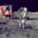 Astronaut salutes beside U.S. flag on the moon. Lunar module visible. Gray surface, blackness of space above.