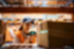 Warehouse worker scans a box with a barcode scanner, wearing an orange vest and blue cap. Shelves with boxes in the blurred background.