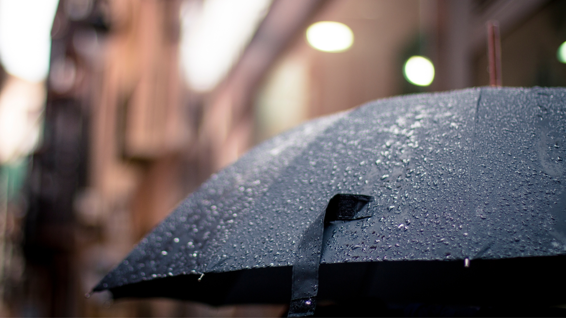 Close-up of a black umbrella with raindrops, set against a blurry cityscape background with soft lights, creating a rainy, melancholic mood.