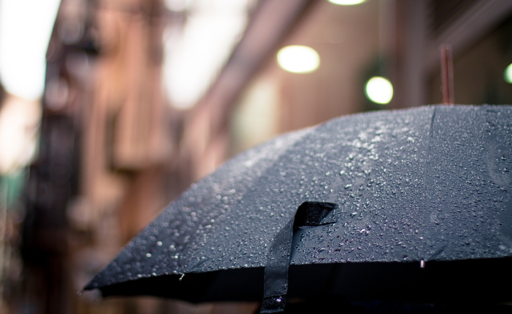 Close-up of a black umbrella with raindrops, against a blurred city street background. Soft lights create a calm, rainy day mood.