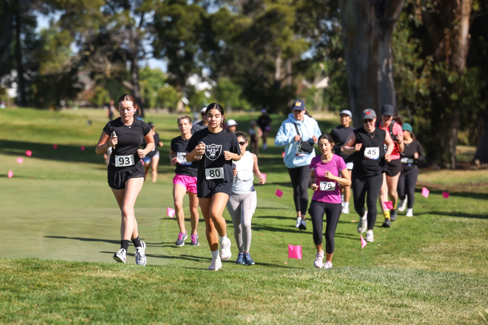A group of runners, mostly women, competing in a race on a grassy path with pink markers.