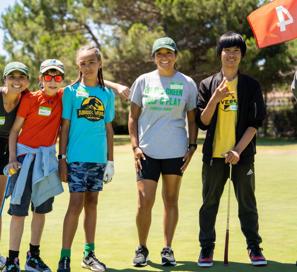 Five kids and adults smiling on a golf course in sunny weather, holding golf clubs and standing together.