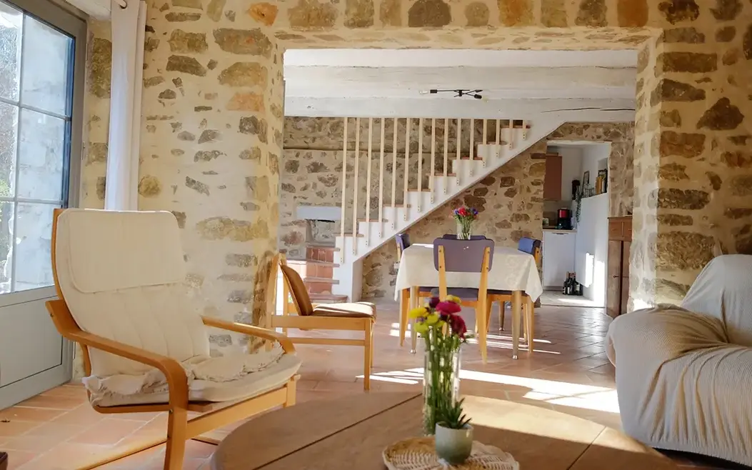 Salon de maison en pierre baignée de soleil avec un bouquet de fleur, fauteuils et chaises de seconde main