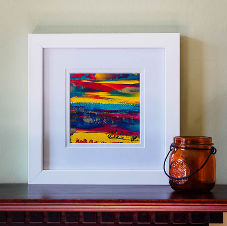 A small bright abstract painting in a white frame next to a glass jar