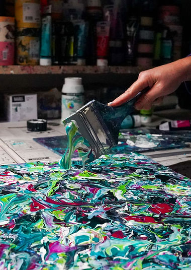 A hand holding a large paint brush dripping with green and white paint over a painted canvas