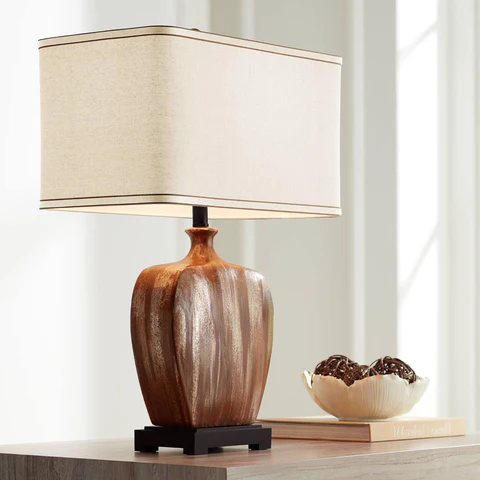 A wooden lamp with a beige shade sits on a table beside a bowl of chocolates and a book. Sunlight filters through a window in the background.