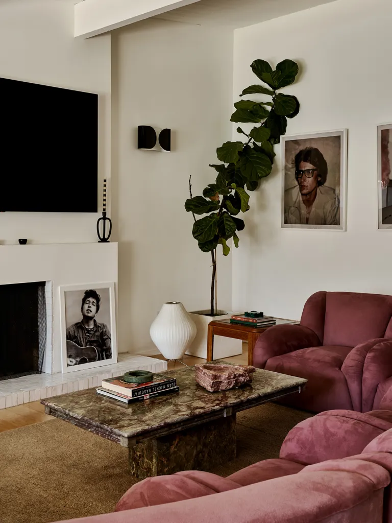 Cozy living room with pink chairs, a marble coffee table holding books, lush green plant, wall art, large TV, and neutral decor.