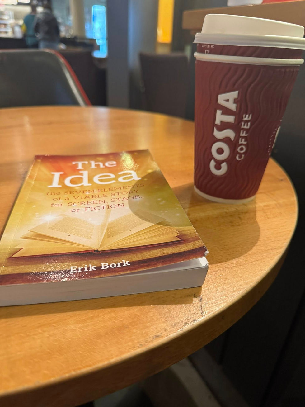 Book titled "The Idea" and a Costa coffee cup on a wooden table. Background shows a coffee shop interior with chairs, evoking a cozy mood.