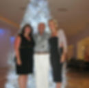 Three people smiling in front of a lit white Christmas tree indoors. Two women wear black dresses, one man in patterned shirt and white pants.
