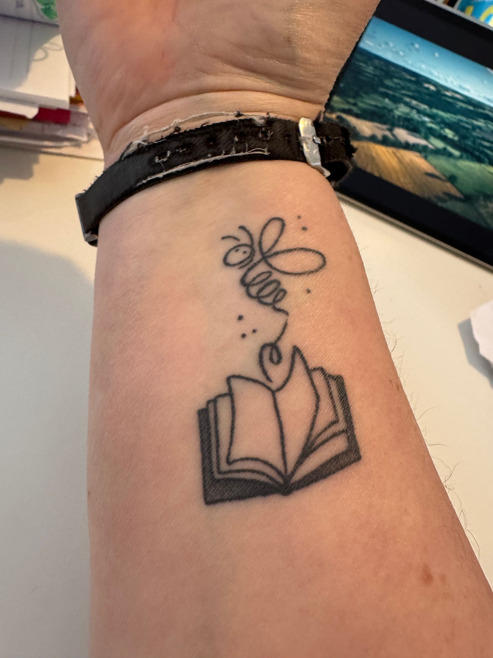 Tattoo of a bee above an open book on a wrist. Background shows a watch band and a blurred image of a landscape on a screen.