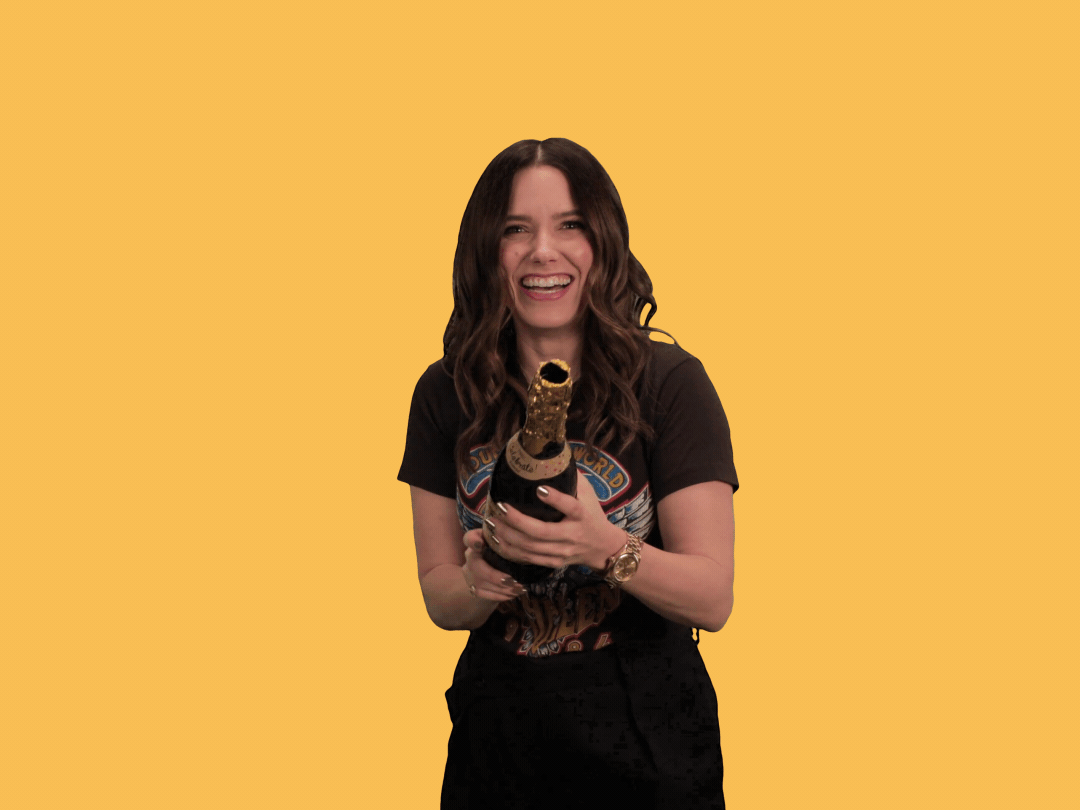 A gif of a happy woman popping a champagne bottle and shooting confetti in celebration