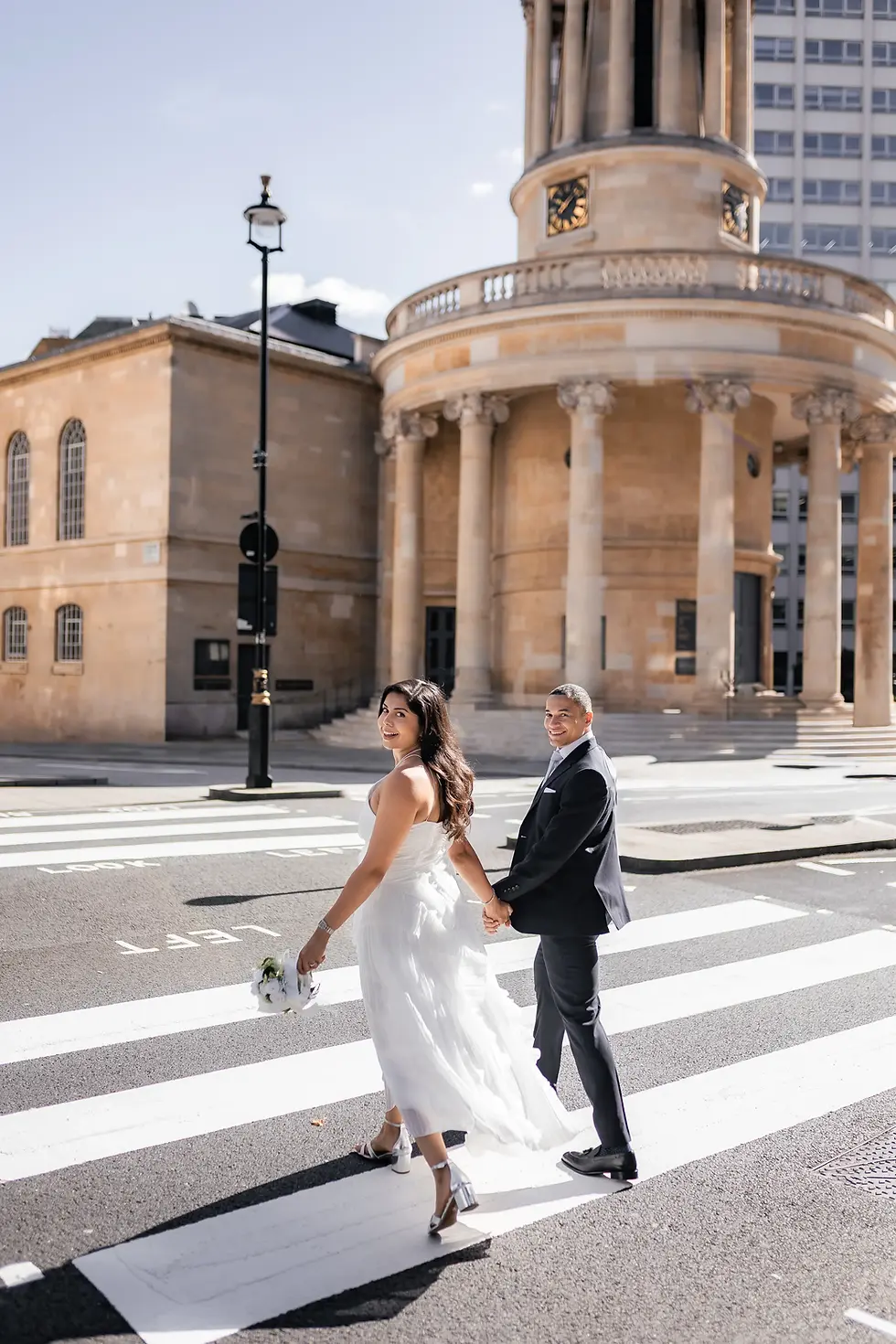 Bride and groom holding hands, smiling while crossing a sunny street. She wears a white dress, and he’s in a dark suit. Historic building behind.