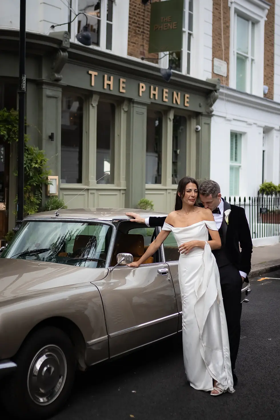 Bride in white dress and groom in black tux pose by vintage car outside "The Phene" pub. Romantic and classic setting.