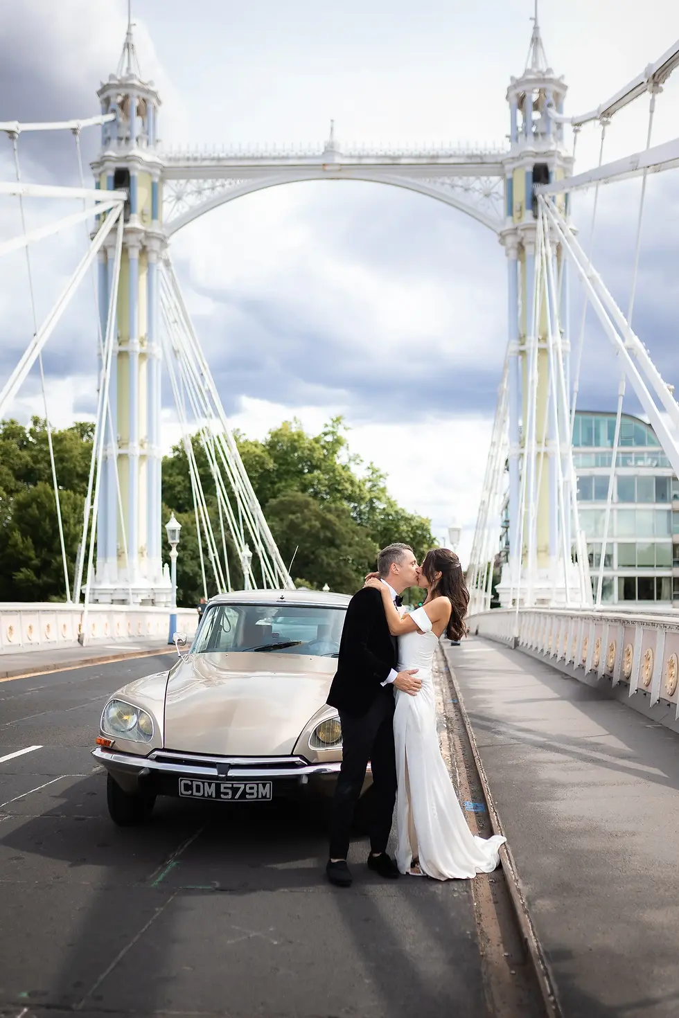 Couple kissing on a bridge beside a vintage car. Overcast sky, trees in background. The woman wears a white dress; the man a dark suit. License plate: CDM 579M.
