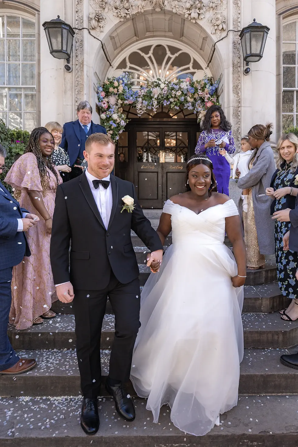 Bride in white gown and groom in black suit exit building, holding hands, surrounded by guests. Flower arch and confetti add festive mood.