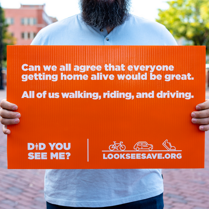 Cody holds a sign that says " Can we all agree that everyone getting home alive would be great. All of us walking, riding, and driving." as part of the Look.See.Save. Campaign.