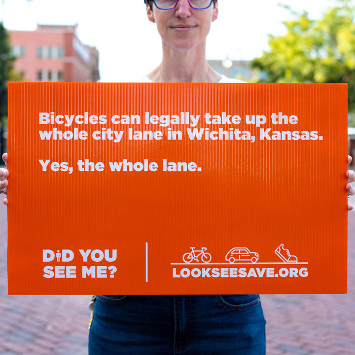 Holly holds a sign that says "Bicycles can legally take up the whole city lane in Wichita, Kansas." as part of the Look.See.Save. Campaign.