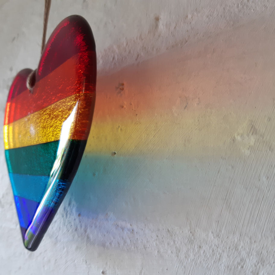 Rainbow glass heart casting colorful light on a white wall, Tam's Glass.