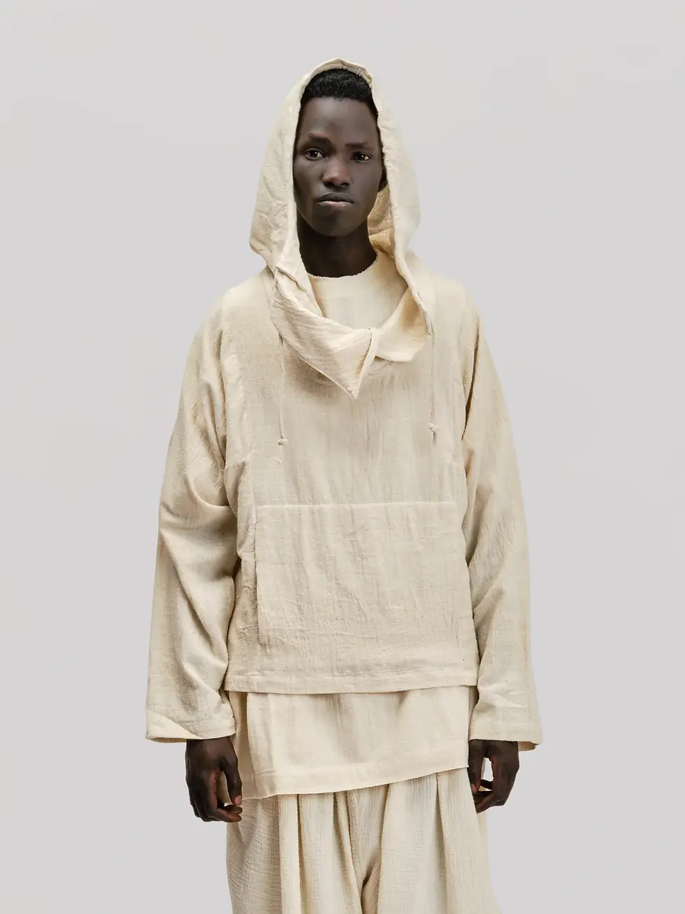 Person in a beige hooded outfit stands against a plain background. The fabric appears textured, adding a minimalist and serene mood.