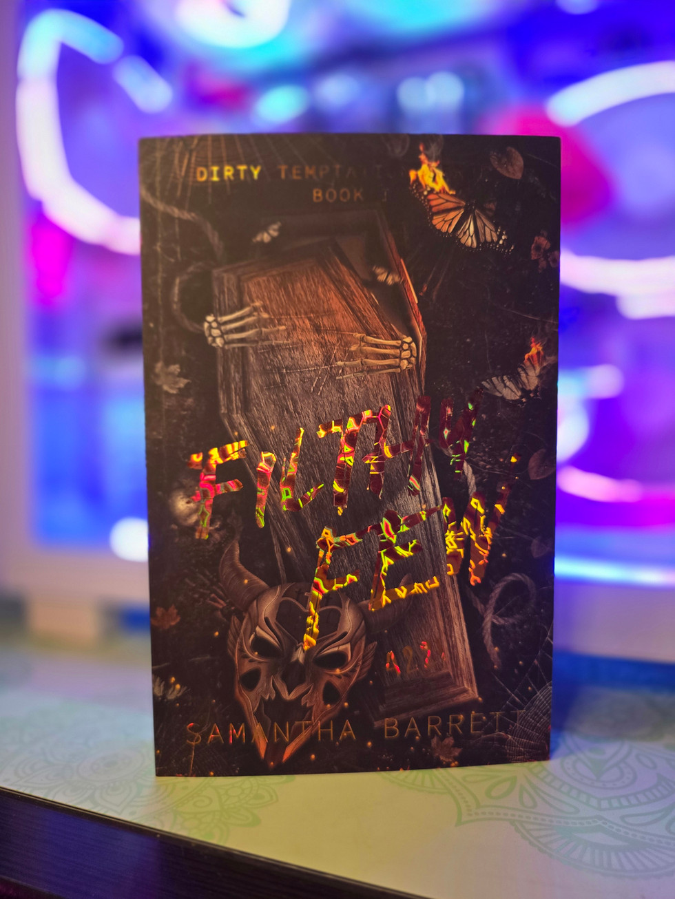 Paperback of Filthy Few by Samantha Barrett, Book I of the Dirty Temptation Series. Dark cover featuring a wooden coffin with skeletal hands, a skull mask, butterflies, and holographic rainbow foil title lettering.