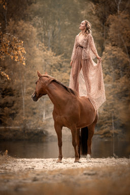 Frau im fließenden Kleid steht auf einem Pferd am See – magischer Moment im Boho-Stil