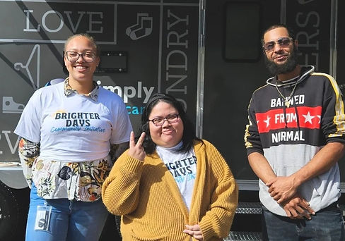 Three young people stand in front of a mobile laundry trailer. The middle person is flashing a peace sign.