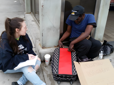 A young woman with a notebook on her lap sits talking with a man who has a physical disability and lives on the streets.