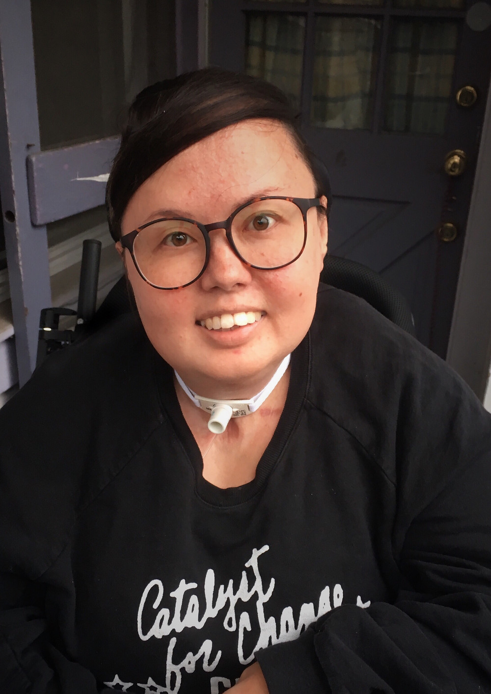 Stacey Milbern has short hair and round glasses. They are smiling at the camera, and wearing a black sweater with white text: Catalyst for Change