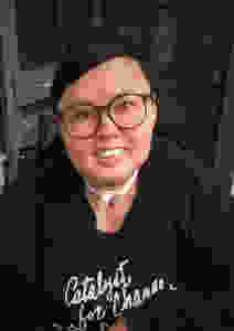 Stacey Milbern has short hair and round glasses. They are smiling at the camera, and wearing a black sweater with white text: Catalyst for Change
