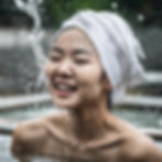 A close-up portrait of a smiling woman with a white towel wrapped around her head, enjoying water splashing on her face. The background shows an outdoor blurred setting, giving a refreshing and natural feel.