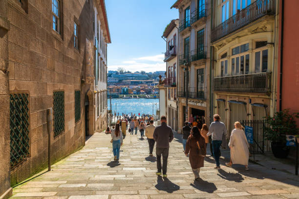 הולכי רגל צועדים ברחוב מרוצף בפורטו לכיוון גדת נהר הדואורו תחת אור השמש.