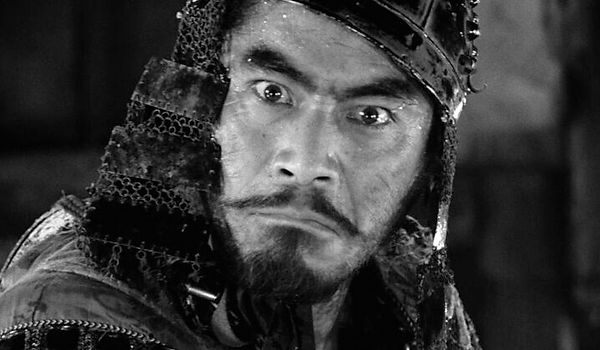 A samurai looks directly at the camera with a mad gaze. He has a helmet on his head and he is seen from the neck up, in a closeup from Akira Kurosawa's cult film The Seven Samurai.