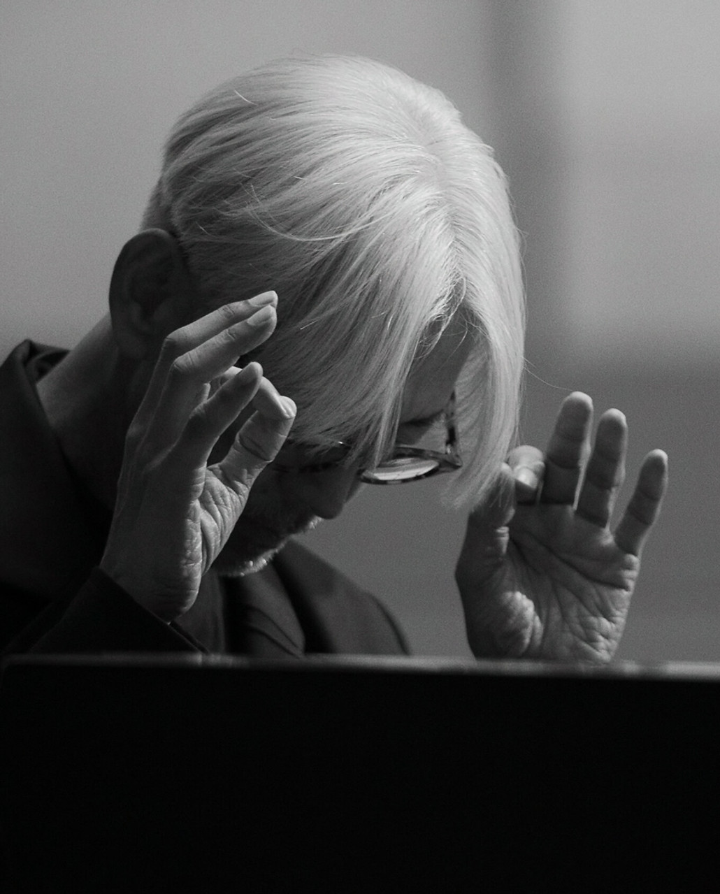 Musician and artist Ryuichi Sakamoto bows his head and raises his hands in front of a black piano, while playing live.