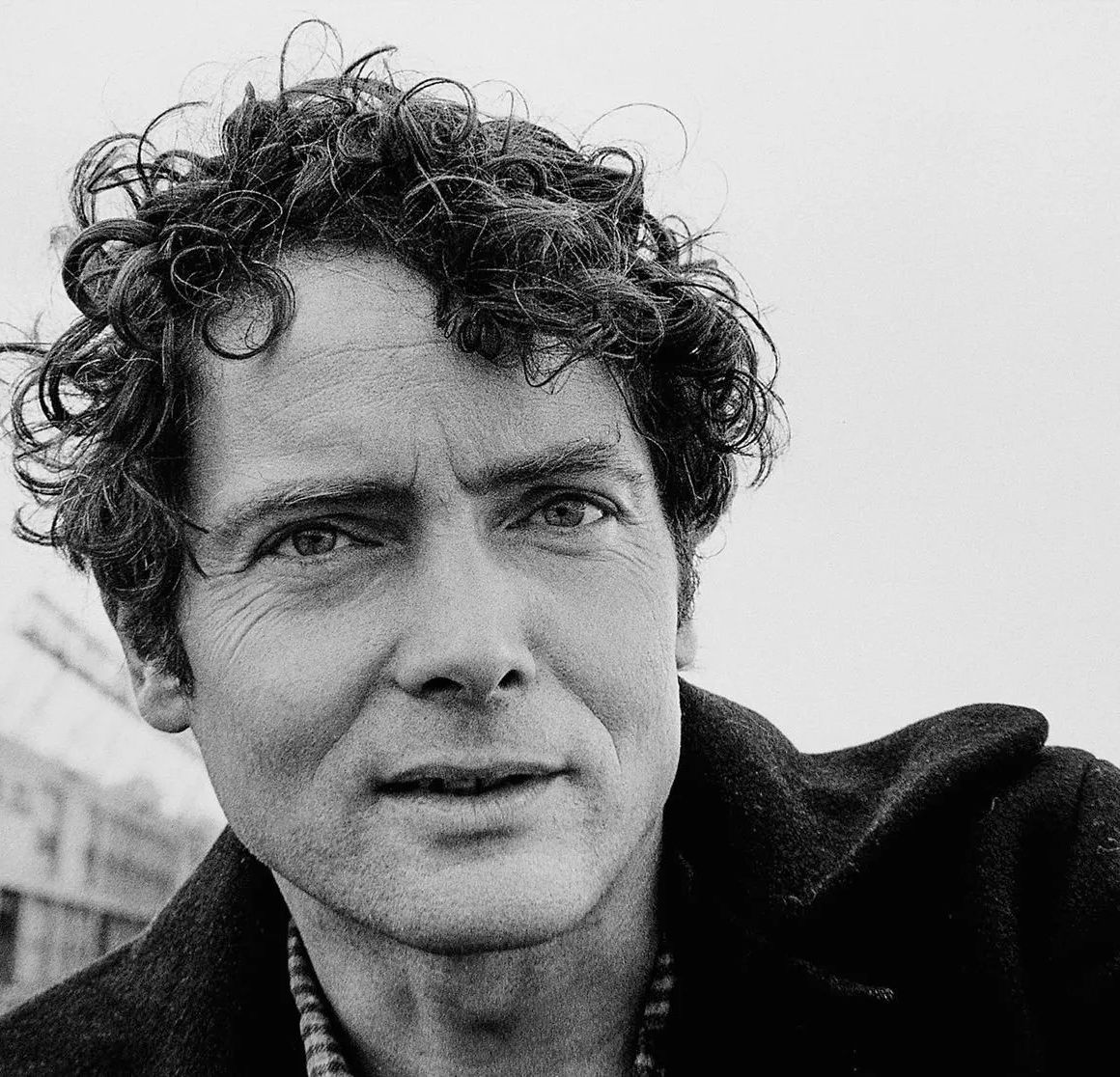 Poet W. S. Merwin looks at the camera while being photographed from the neck up. His hair is short and wavy, and he is wearing a black coat.