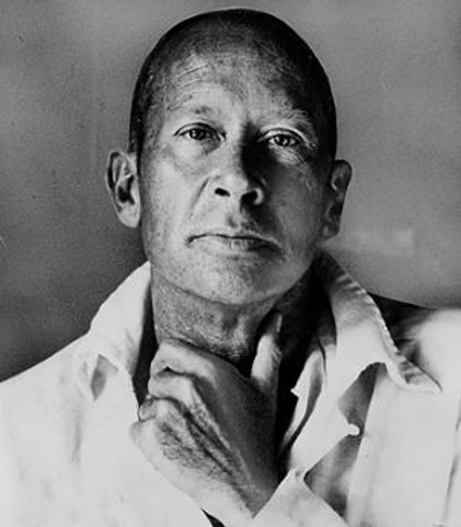 Poet E. E. Cummings looks directly at the camera while being photographed in black and white. He is wearing a white shirt, and his left hand is on his neck.