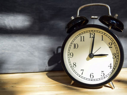 Black vintage alarm clock on a wooden table, showing 3:00, against a dark blurred background.