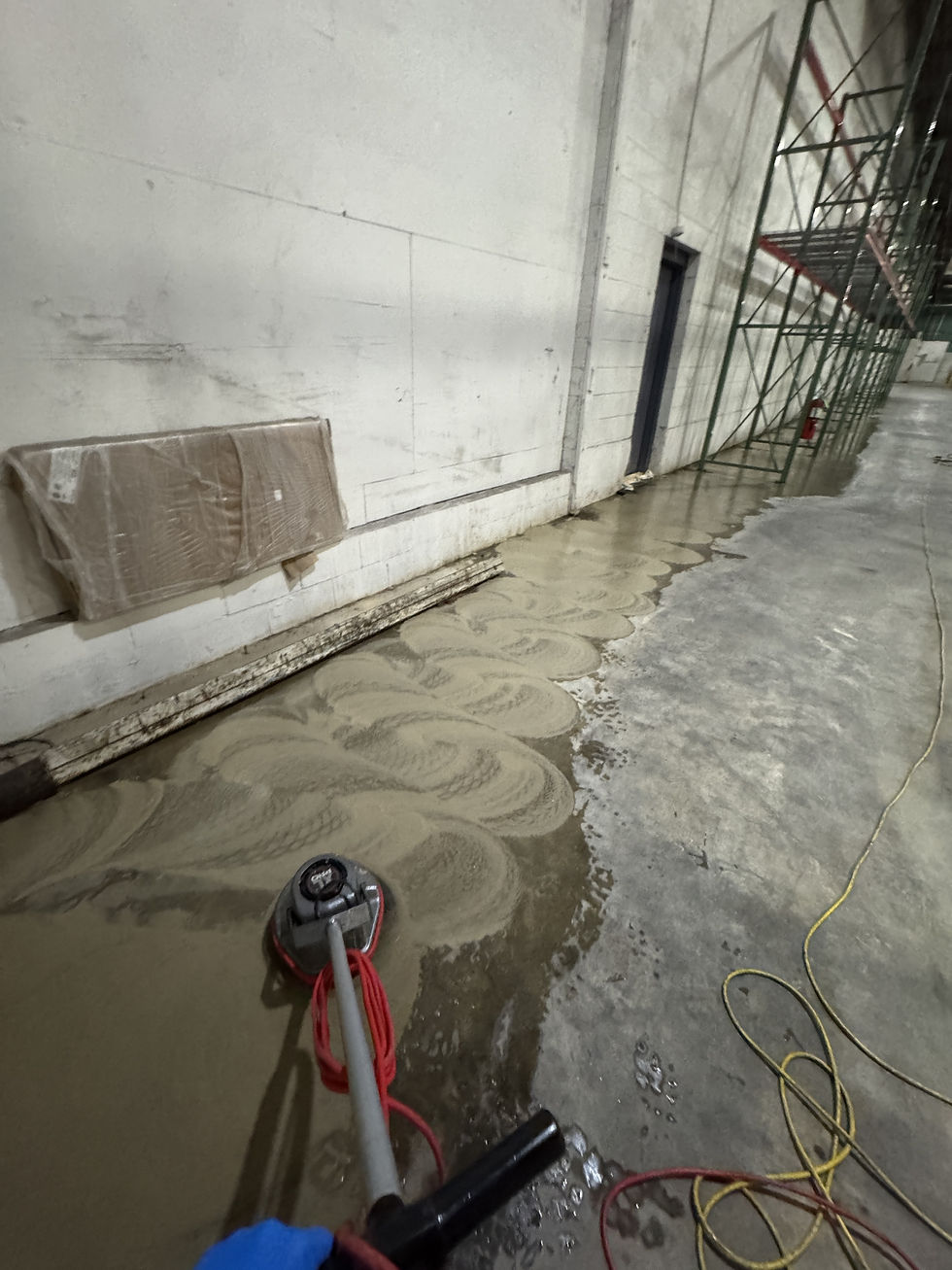 Swing floor machine scrubbing along a warehouse wall, with brown slurry of lifted dirt beside a cleaner strip of concrete.